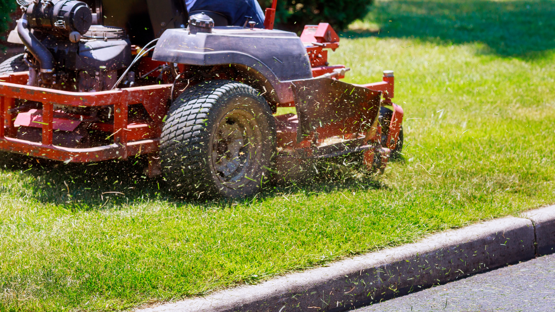 A red riding lawnmower cuts grass along a curb on a sunny day, with clippings flying.
