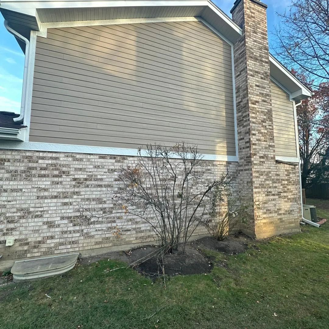 Side of a brick house with a chimney and light brown siding. Bare bush in front.