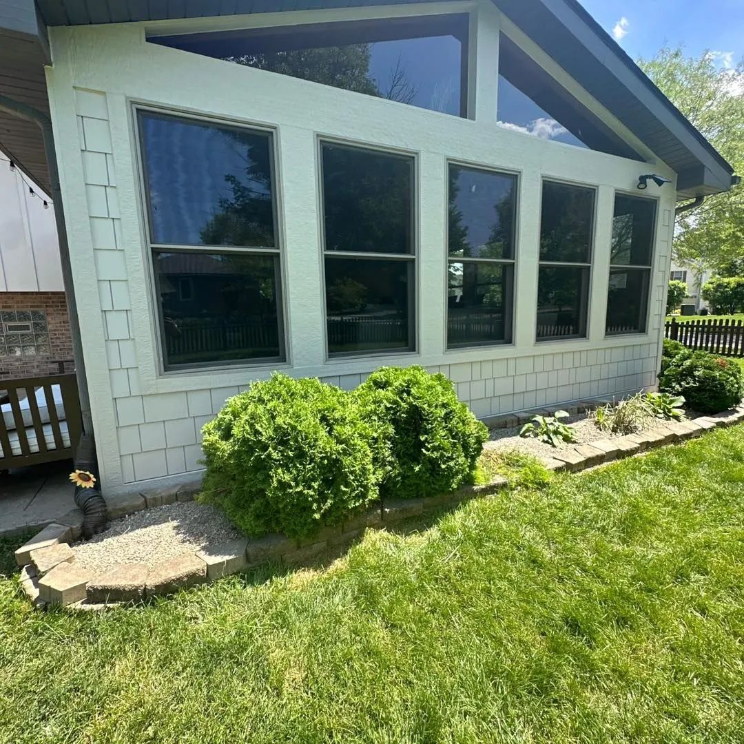 A light-colored sunroom with dark-tinted windows and two green bushes in front. The setti