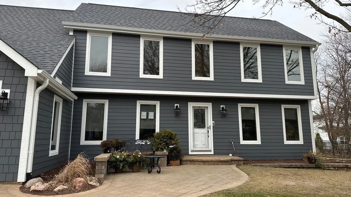 A two-story house with dark gray horizontal siding, a white front door, and a paved stone walkway leading to the entrance.