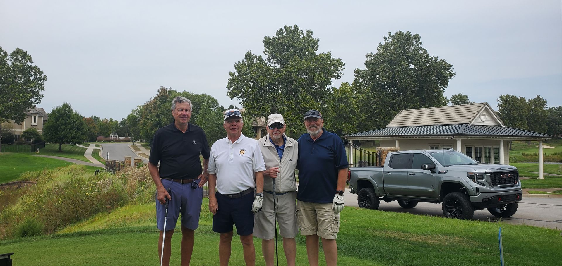 Four men pose for a photo on a golf course. A gray truck is parked nearby, trees, and a cloudy sky in the background.