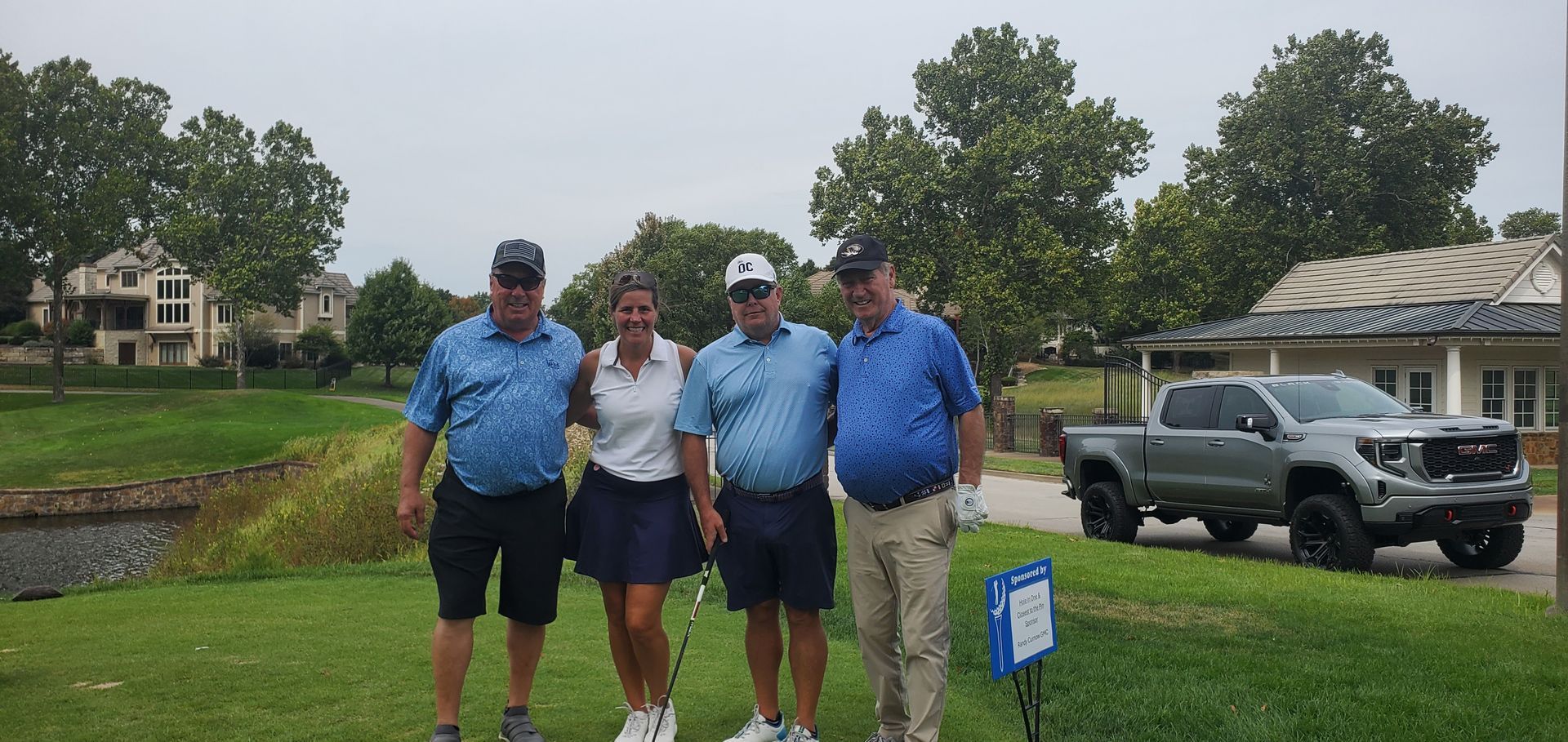 Four golfers standing on the green, smiling. Trees, houses, and a truck are visible in the background.