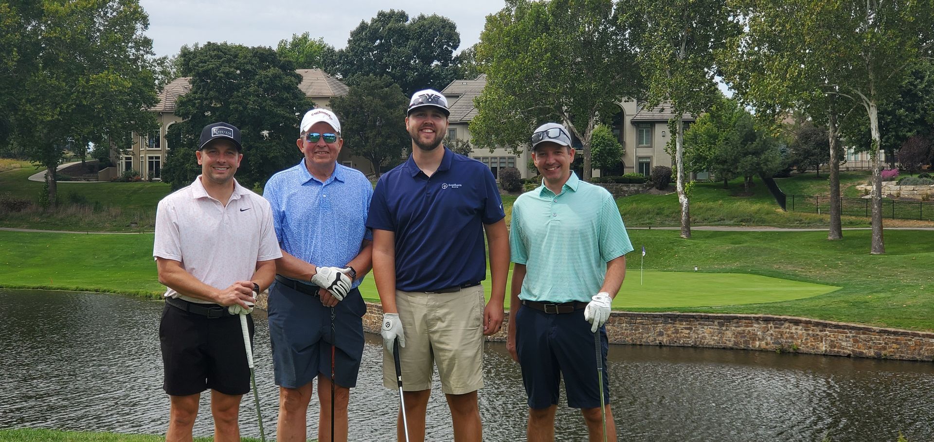 Four men on a golf course, standing near water. All are wearing hats and holding golf clubs.