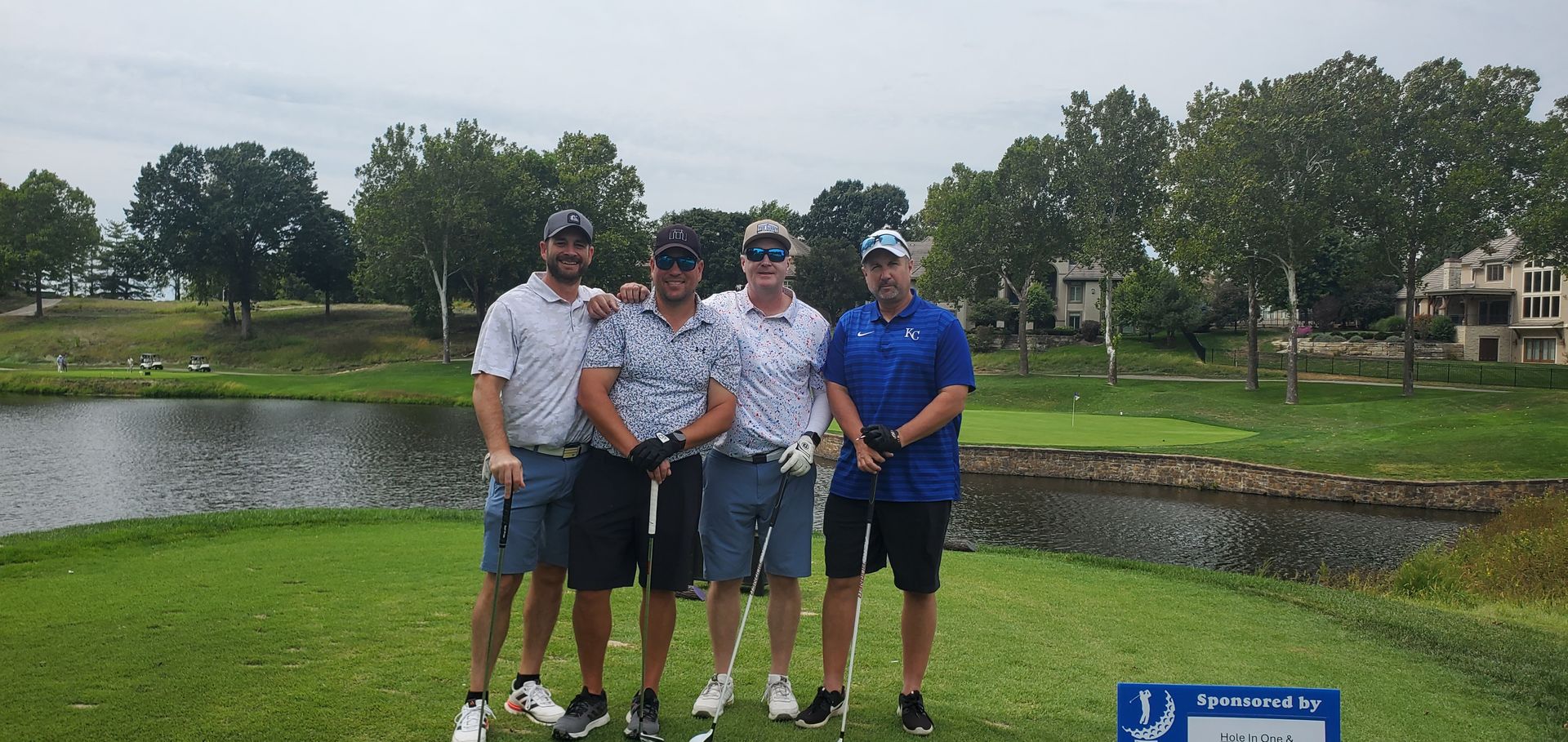 Four men pose for a photo on a golf course. They are holding golf clubs. A lake is in the background.