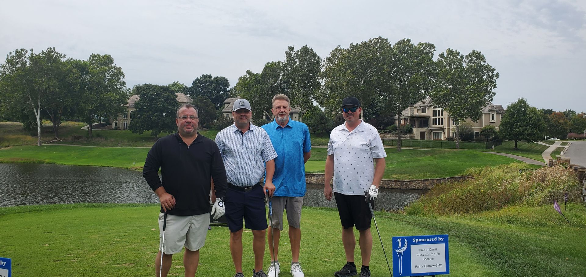Four men stand on a golf course, posing for a photo. They're near a body of water and trees in a golf course setting.