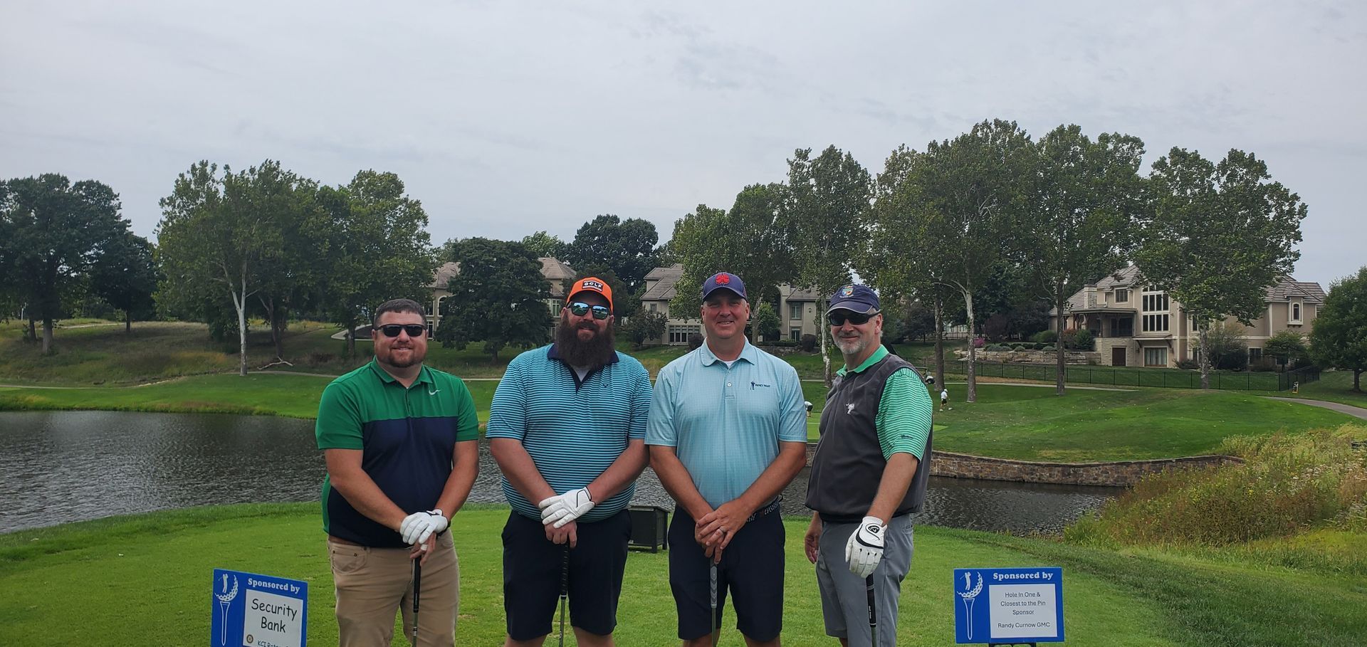 Four men stand on a golf course, holding golf clubs. They are smiling. Overcast day.