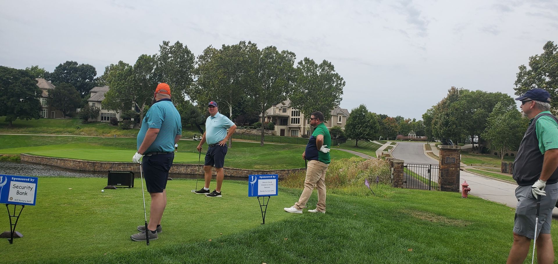 Four golfers stand on a tee box on a cloudy day, preparing to hit.