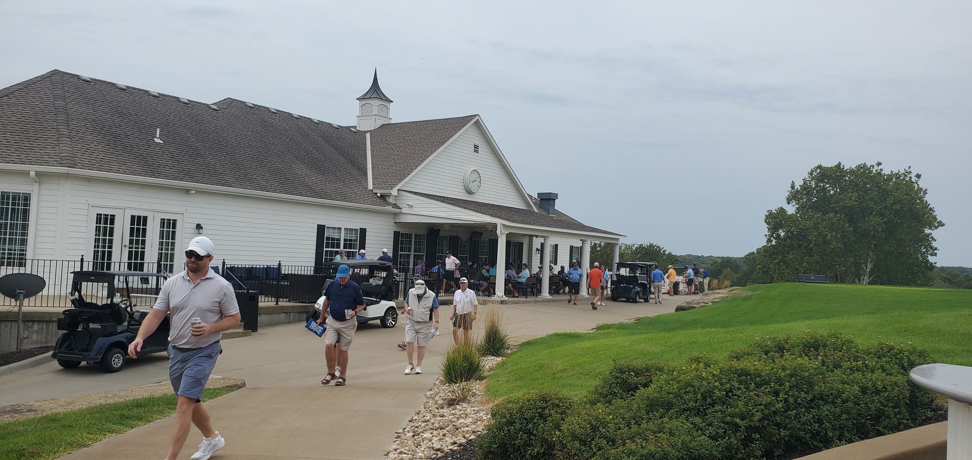 People walking near a white golf clubhouse with a dark roof. Green grass and golf carts are present. Overcast sky.