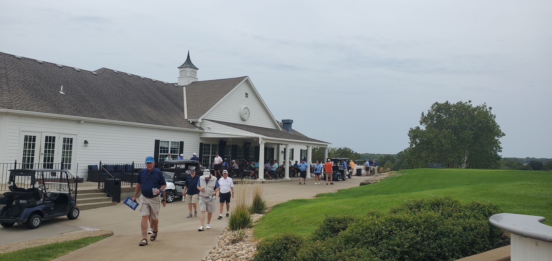 Golfers walking toward a white clubhouse on a cloudy day. Golf carts are parked outside on the green lawn.