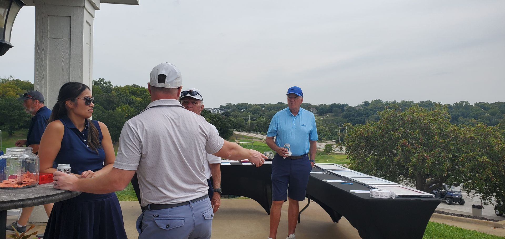 People at an outdoor event, some are talking by a table with paperwork, overlooking a golf course.