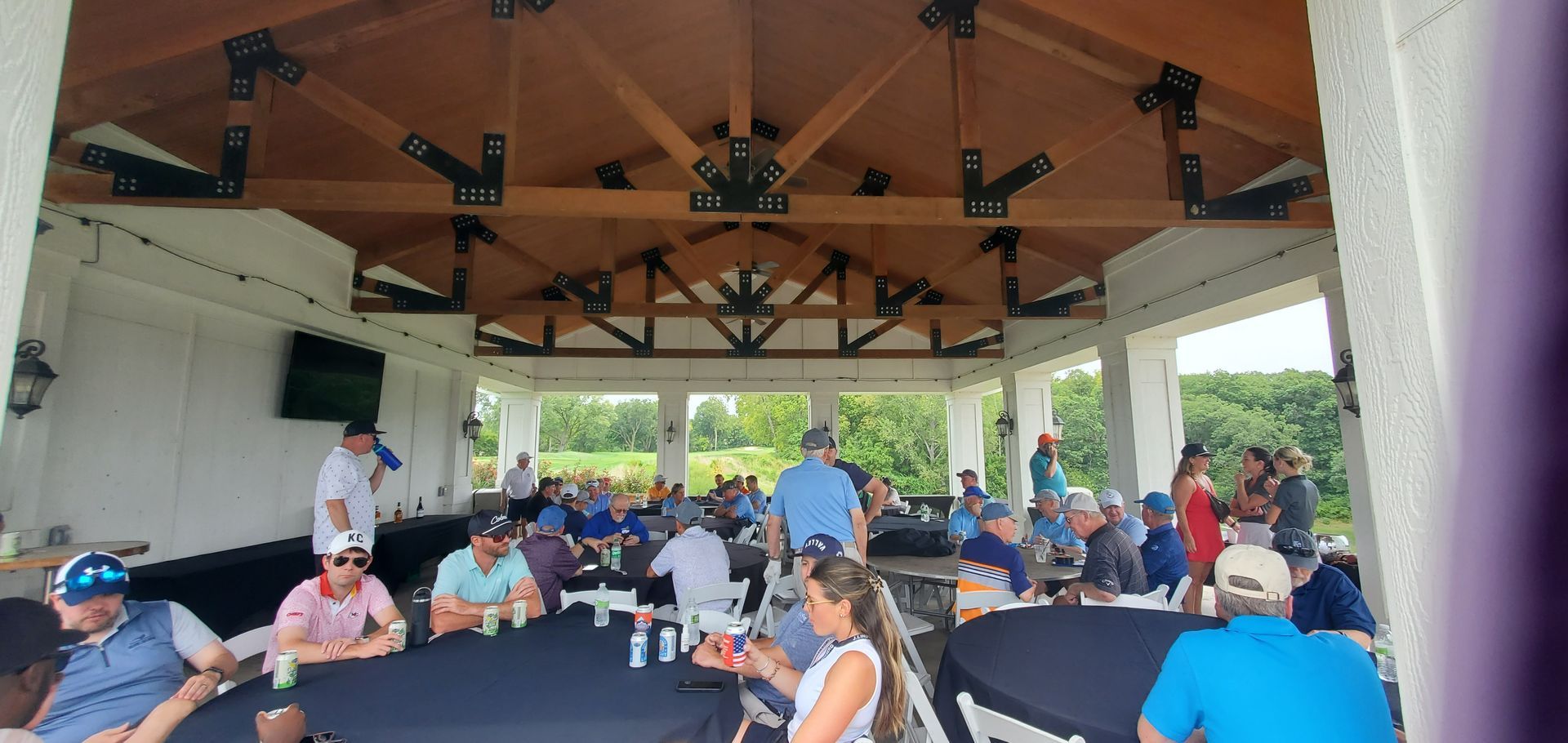 Group of people gathered at an outdoor event under a pavilion. They are seated at tables. The setting is green.