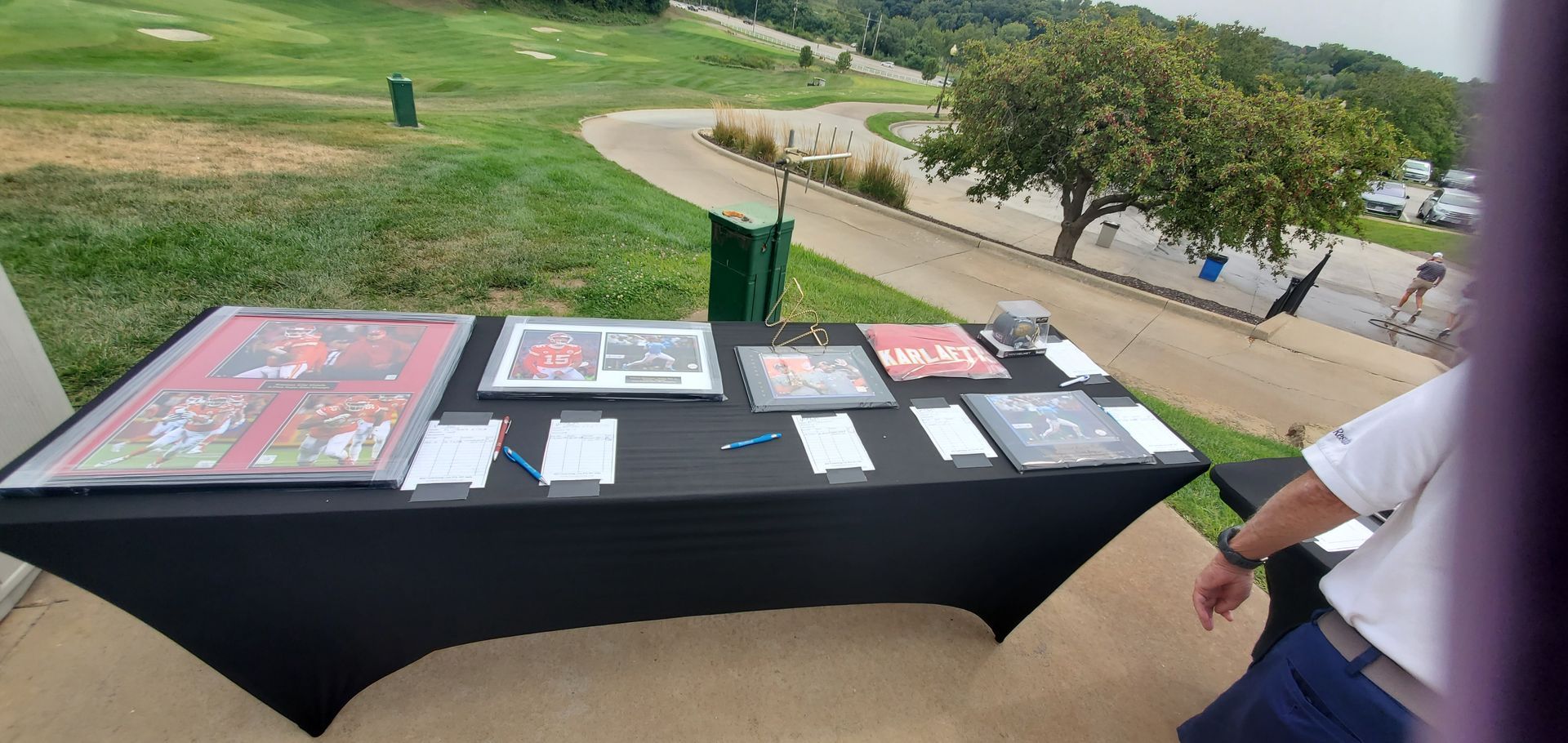 A table with framed items and paperwork, outside on a golf course with a person standing nearby.