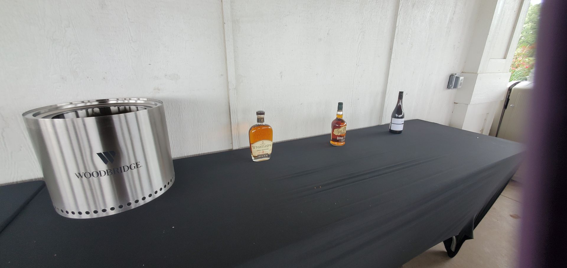 A stainless steel fire pit and bottles of alcohol sit on a black-covered table outdoors.