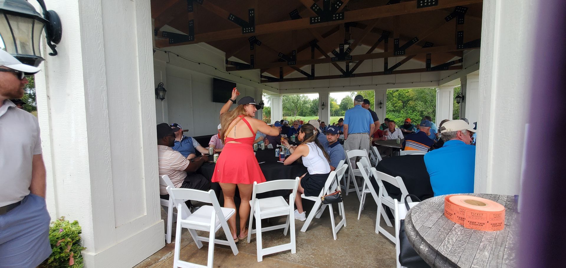 Woman in red dress gestures near a table, pavilion setting, people seated around tables.