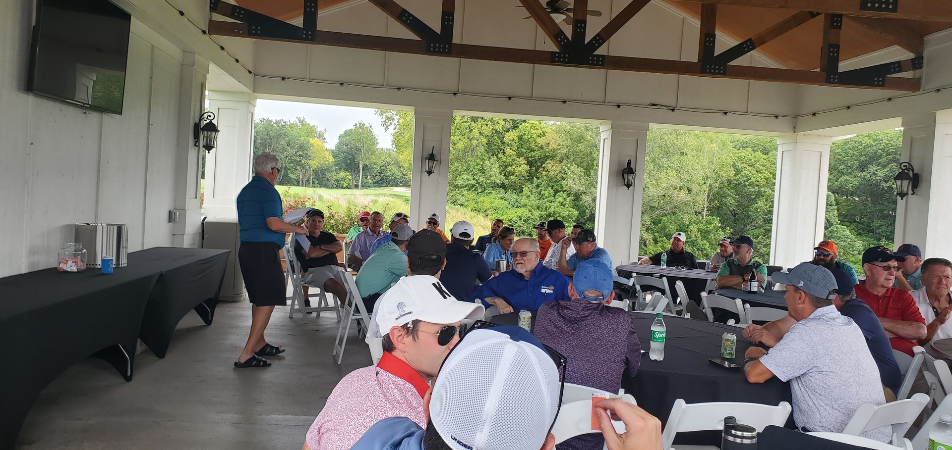 A man gives a presentation to a group of people seated at tables in an outdoor pavilion.
