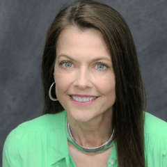 Woman with long brown hair smiling; wearing a green shirt, silver necklace and hoop earrings, against a gray background.