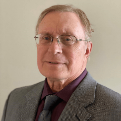Older man with glasses, wearing a suit and purple shirt, smiling slightly.