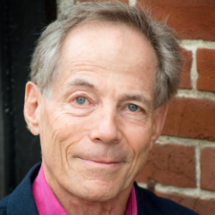Mature man, light eyes, pink shirt, navy blazer, smiles, stands before a brick wall.