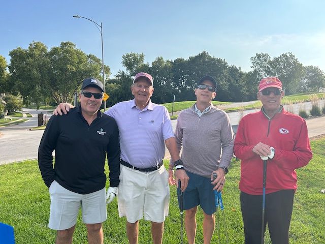 Four men smiling, posing for a photo on a golf course. One in a red Chiefs jacket, others in golf attire.