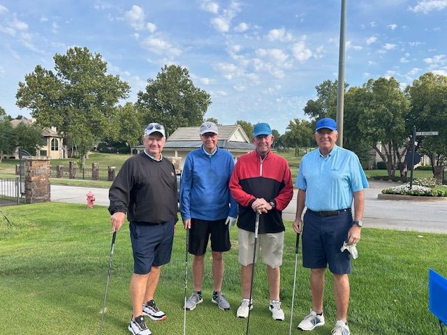 Four men in golf attire on a green, posing with clubs. Overcast day, trees, houses visible.