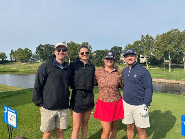 Four people pose on a golf course. Two men, two women, wearing golf attire, smiling. Green grass, blue sky, and a pond.