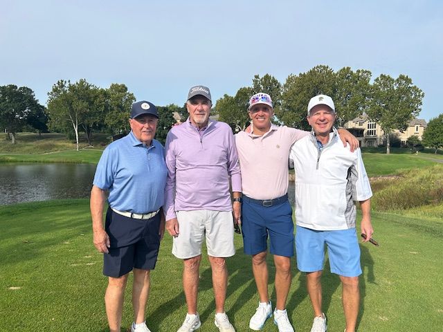 Four men smiling on a golf course. They wear golf attire, posing near a lake.