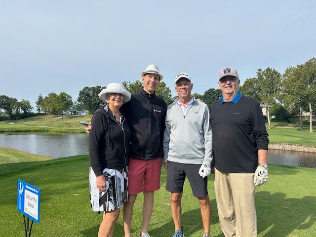 Four people pose on a golf course. Two men and two women wearing hats and casual attire stand on green grass near a pond.
