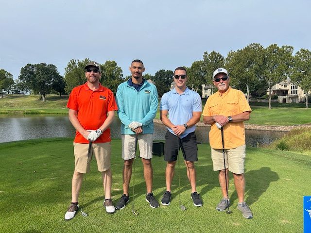 Four men on a golf course. One in orange shirt, three holding clubs, standing in front of a lake.