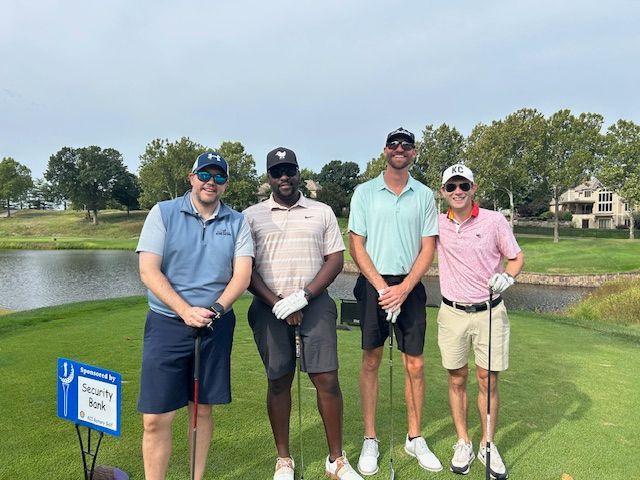 Four men on a golf course, smiling, holding clubs. Green grass, pond, and trees in the background.