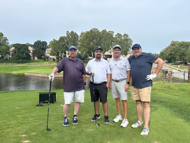Four men standing on a golf course, posing with clubs; green grass, pond, and trees in background.