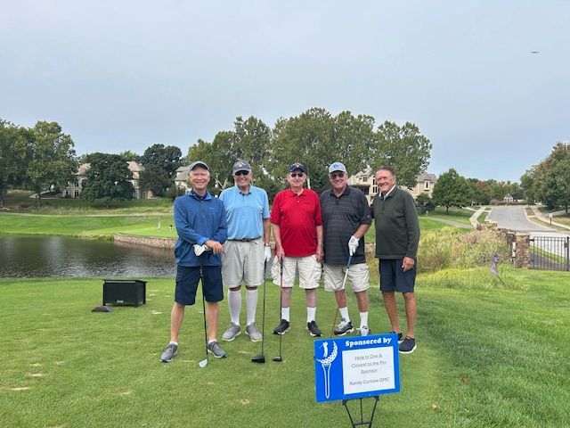 Five men pose on a golf course; one holds a club. A lake and houses are in the background.