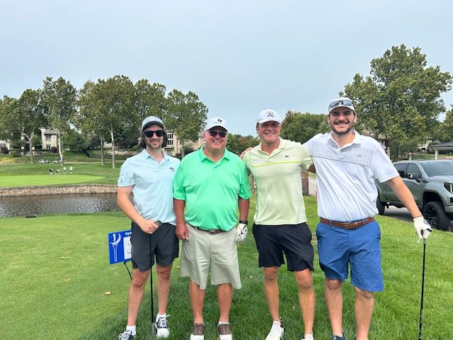 Four men smiling, posing on a golf course. Green grass, water, and trees in the background.