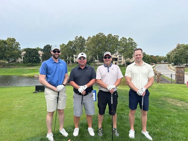 Four men on a golf course posing with clubs; green grass, water, and trees in background.