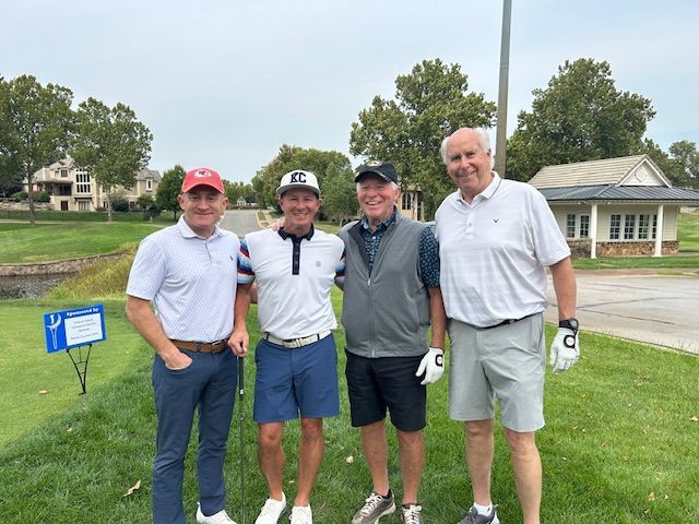Four men pose on a golf course: wearing golf attire, smiling, and standing near a sign and building.