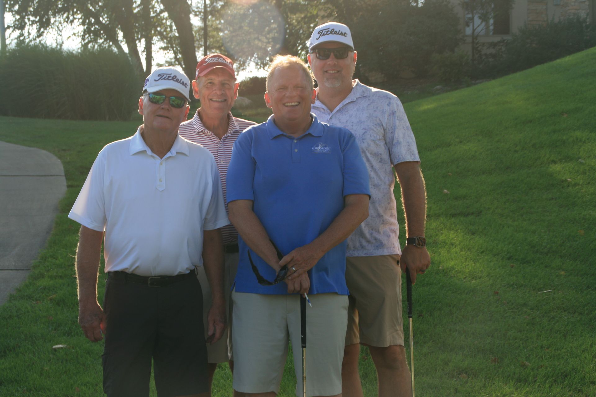 Four men in golf attire pose on a green. All wear hats, smiles, and hold golf clubs. Sunny outdoor setting.
