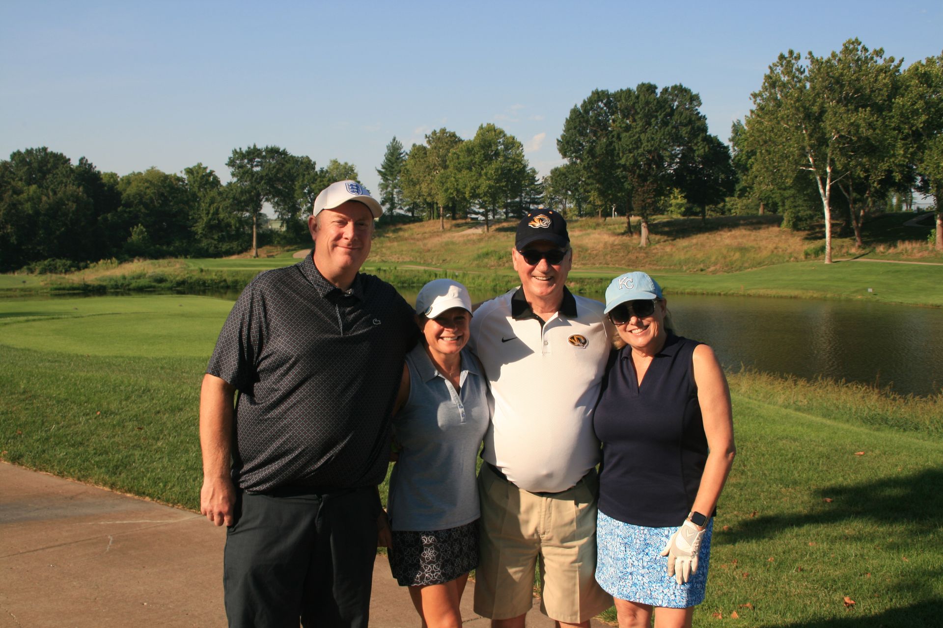 Four people smiling on a golf course, trees and a pond in the background.