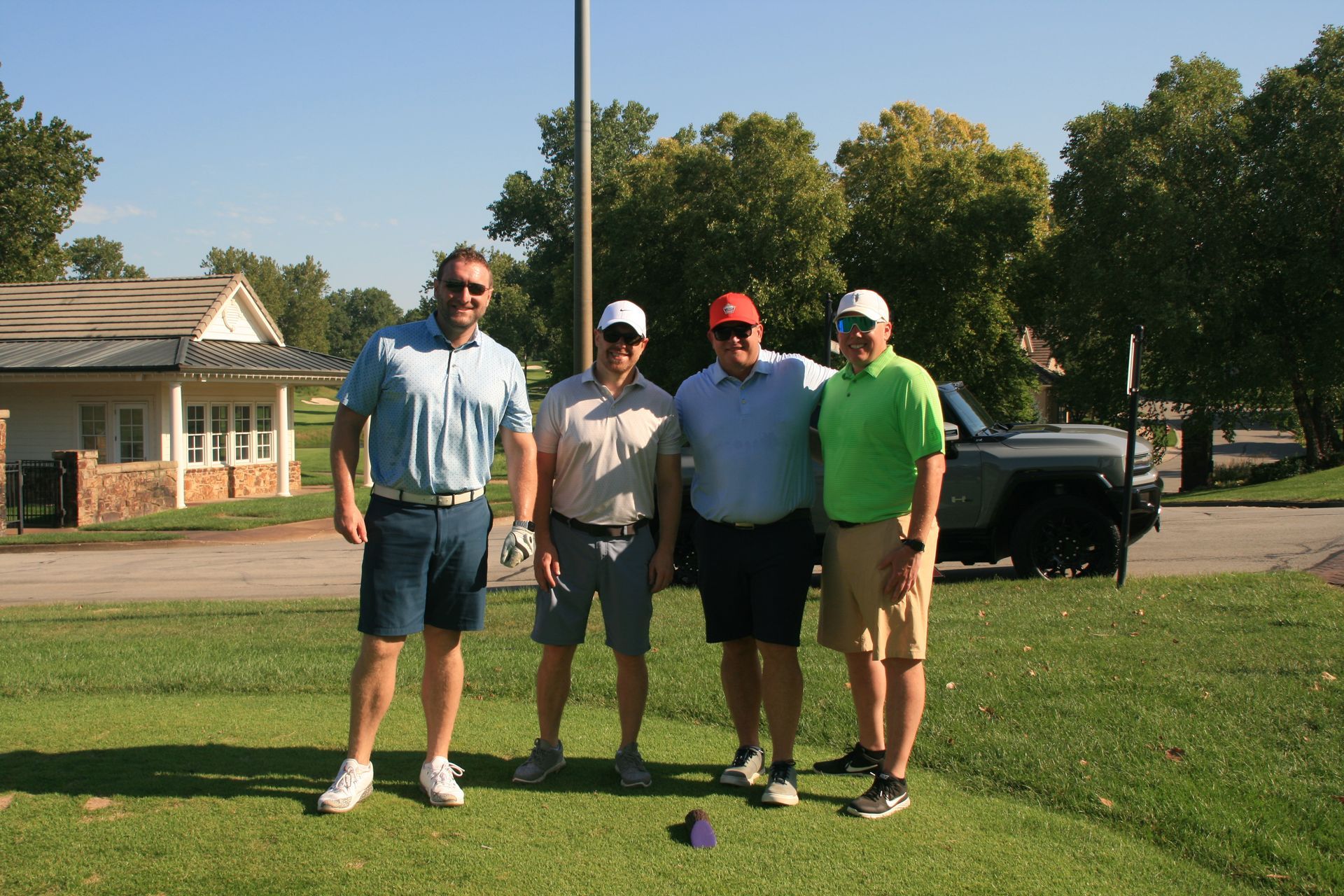Four men in golf attire standing on a green, smiling, near a building and a car.