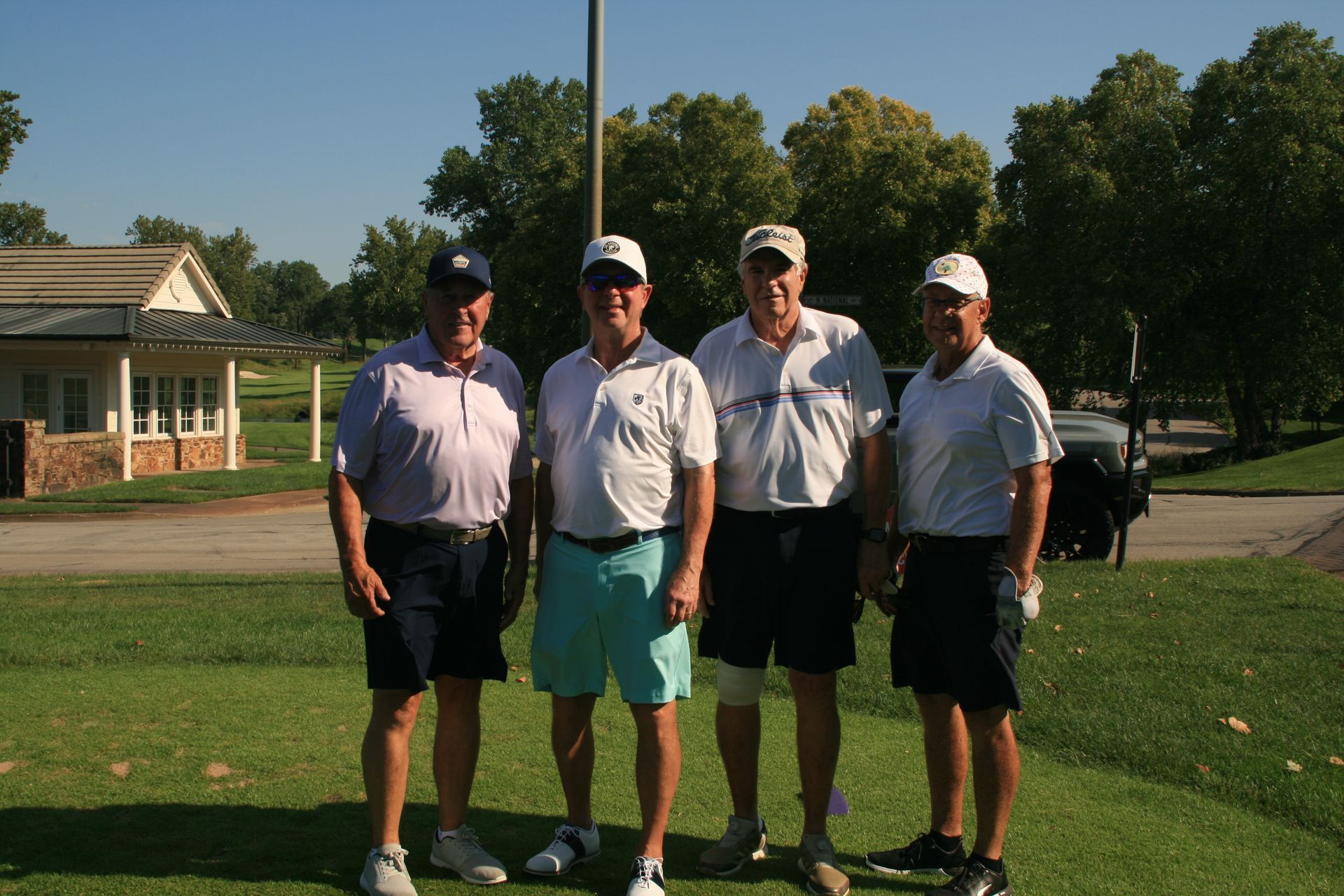 Four men in golf attire pose on a green, sunny day. A golf cart and clubhouse are visible.