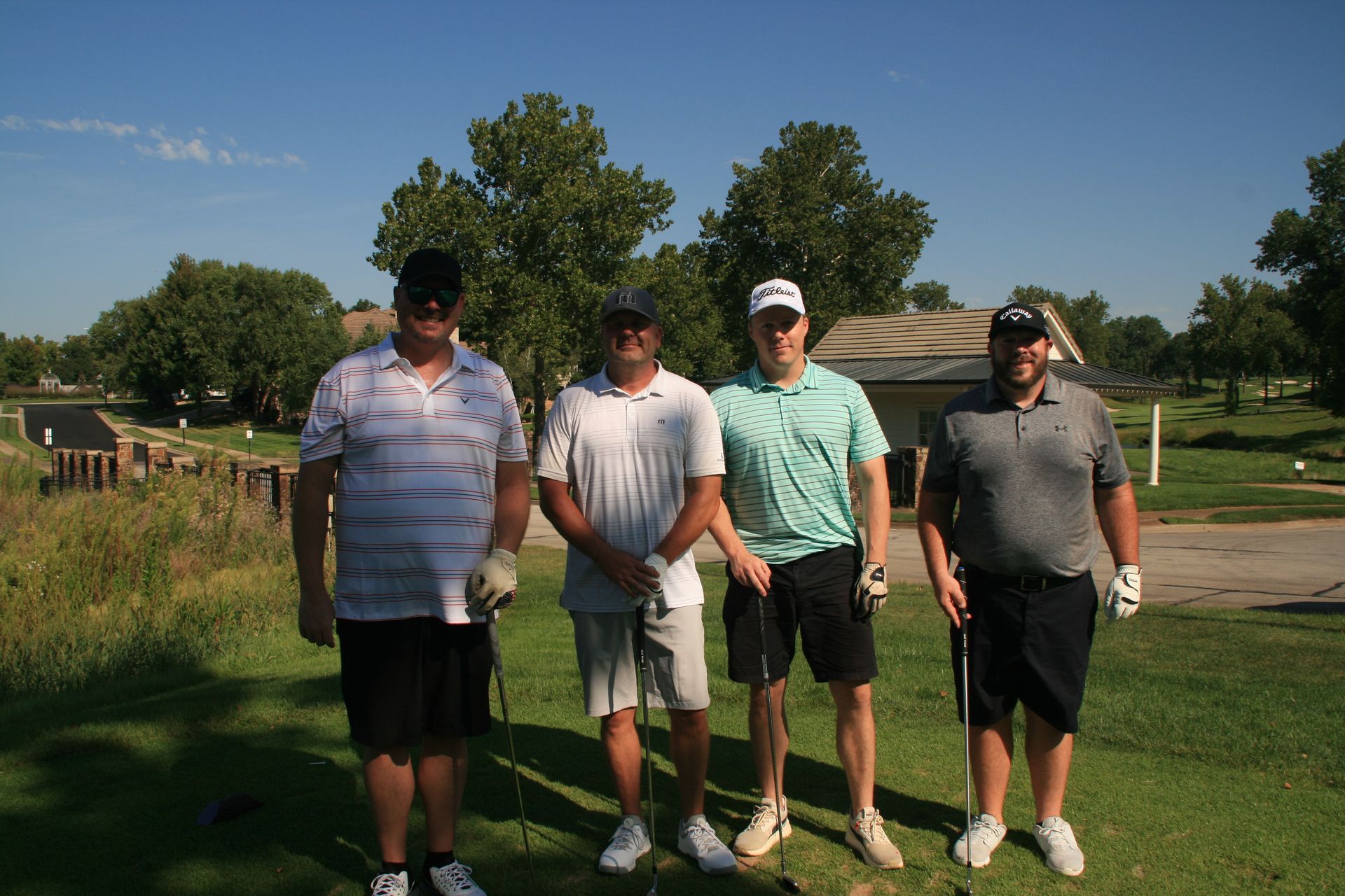 Four men smiling, holding golf clubs on a green, sunny day.