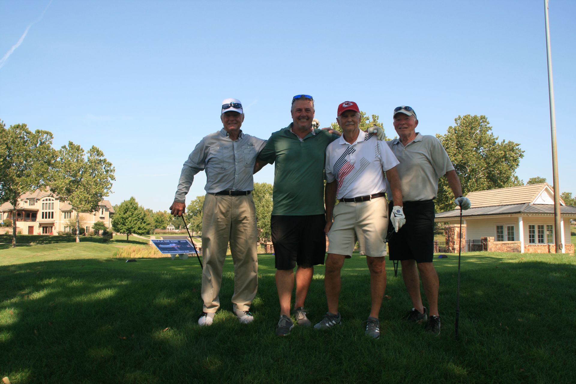 Four golfers posing on a green, sunny day. One has arm around another. Houses and trees in the background.