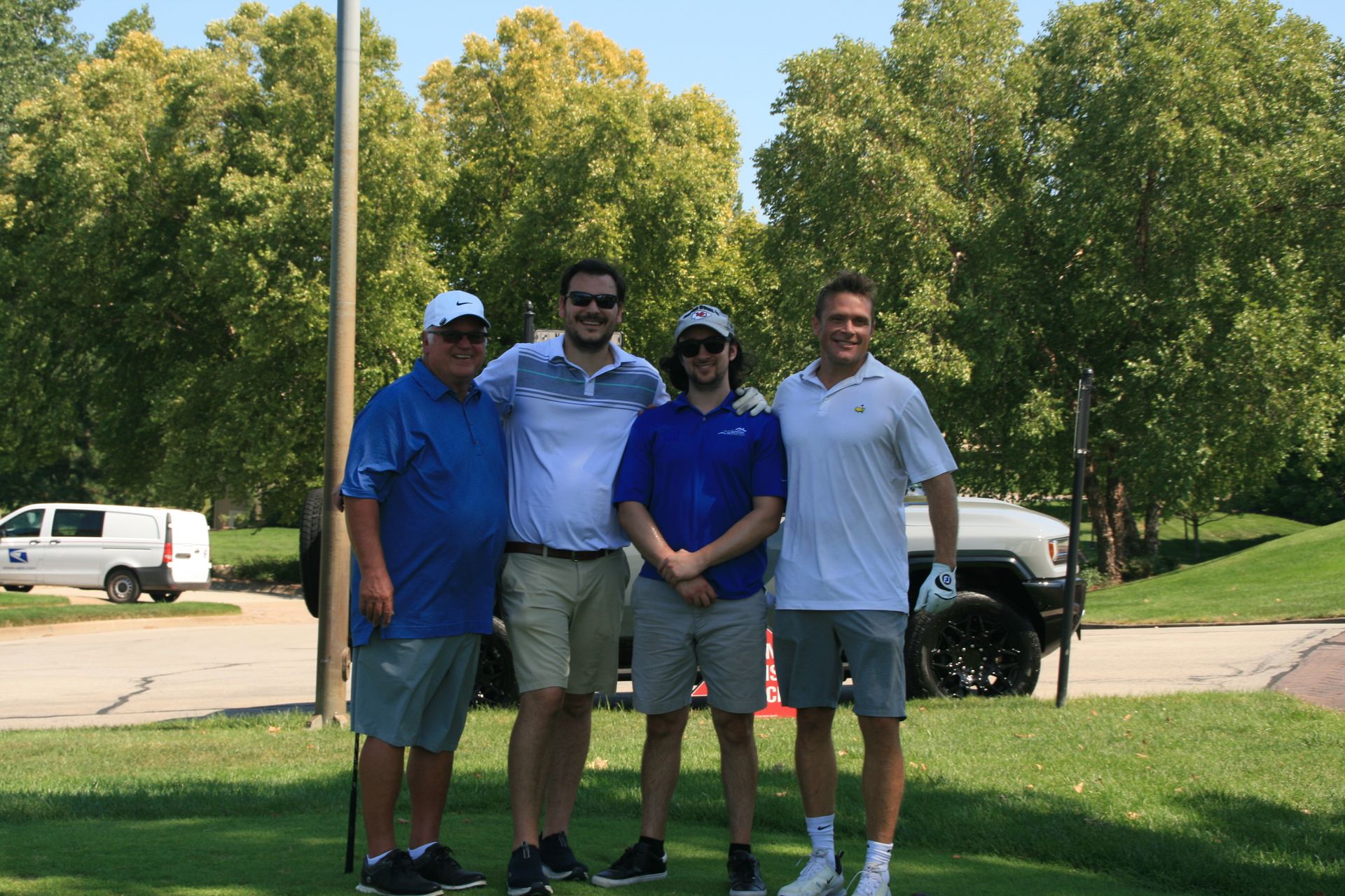Four men in golf attire, posing on a green with a truck and trees in the background.