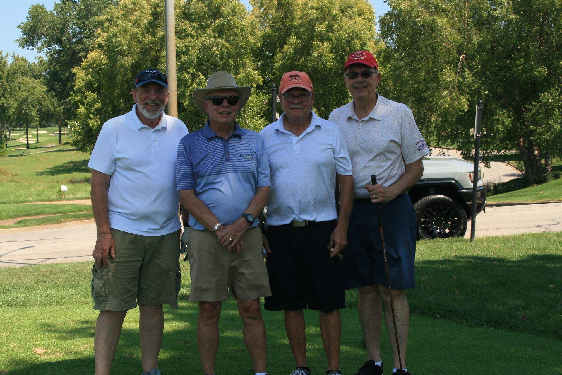Four men pose for a photo on a sunny golf course. They wear golf attire; hats, shorts, and polo shirts.