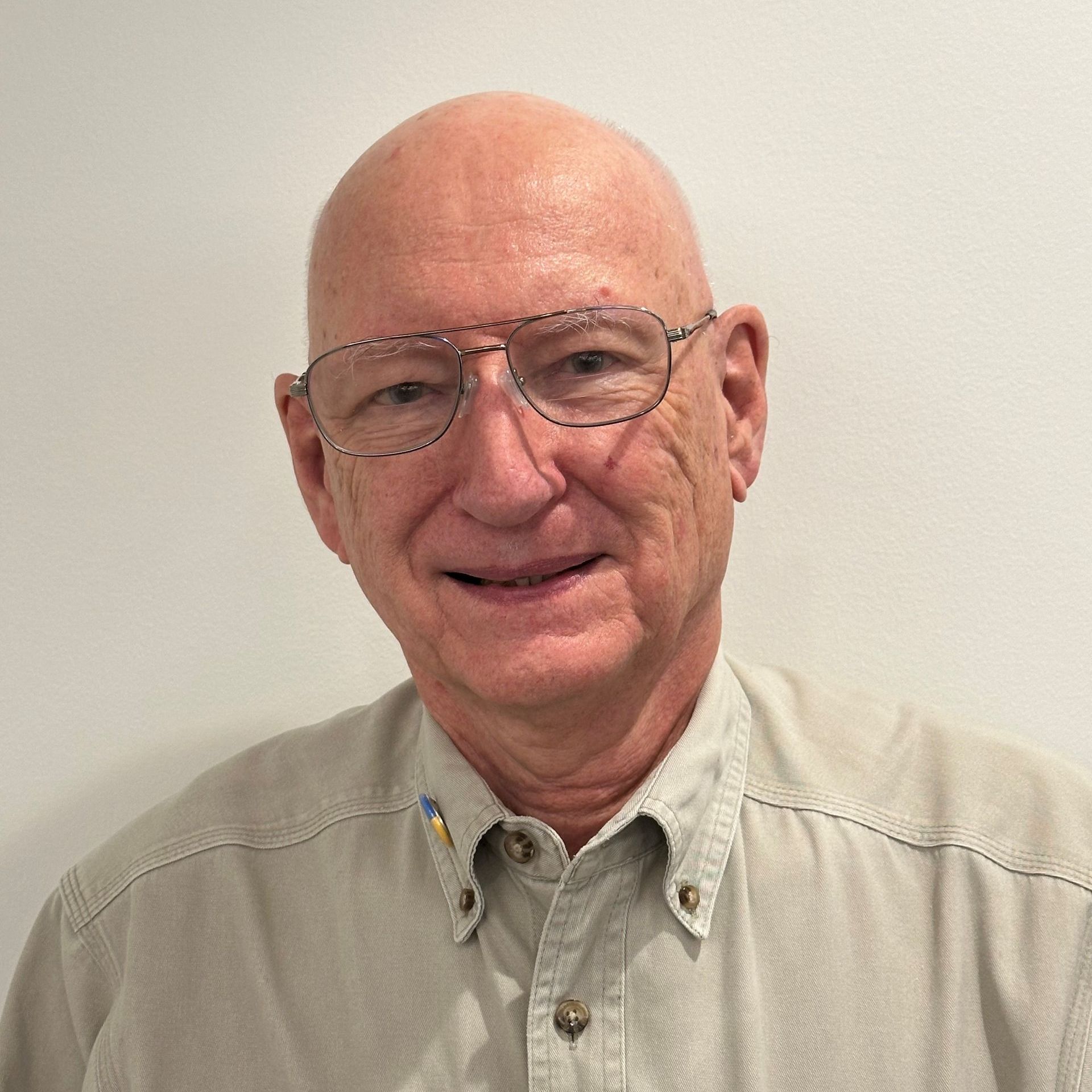 Bald man with glasses smiles, wearing a tan button-down shirt, in front of a white wall.