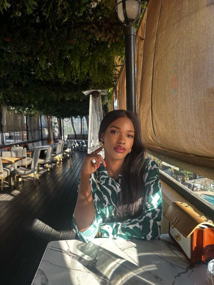 Sophie Namugga's image. Woman seated at a table in a restaurant, wearing green patterned shirt, with long hair and bright sunlight.
