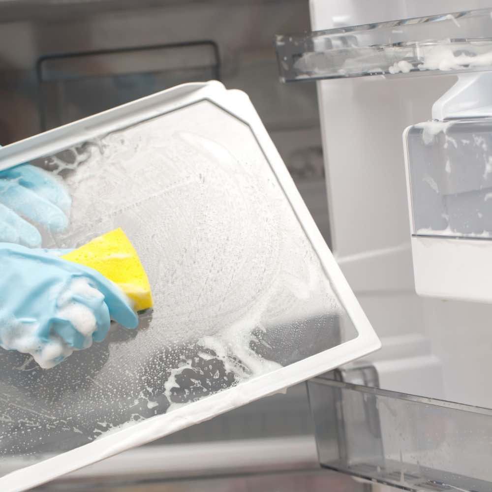 Person wearing blue gloves cleaning a clear refrigerator shelf with a yellow sponge and suds.