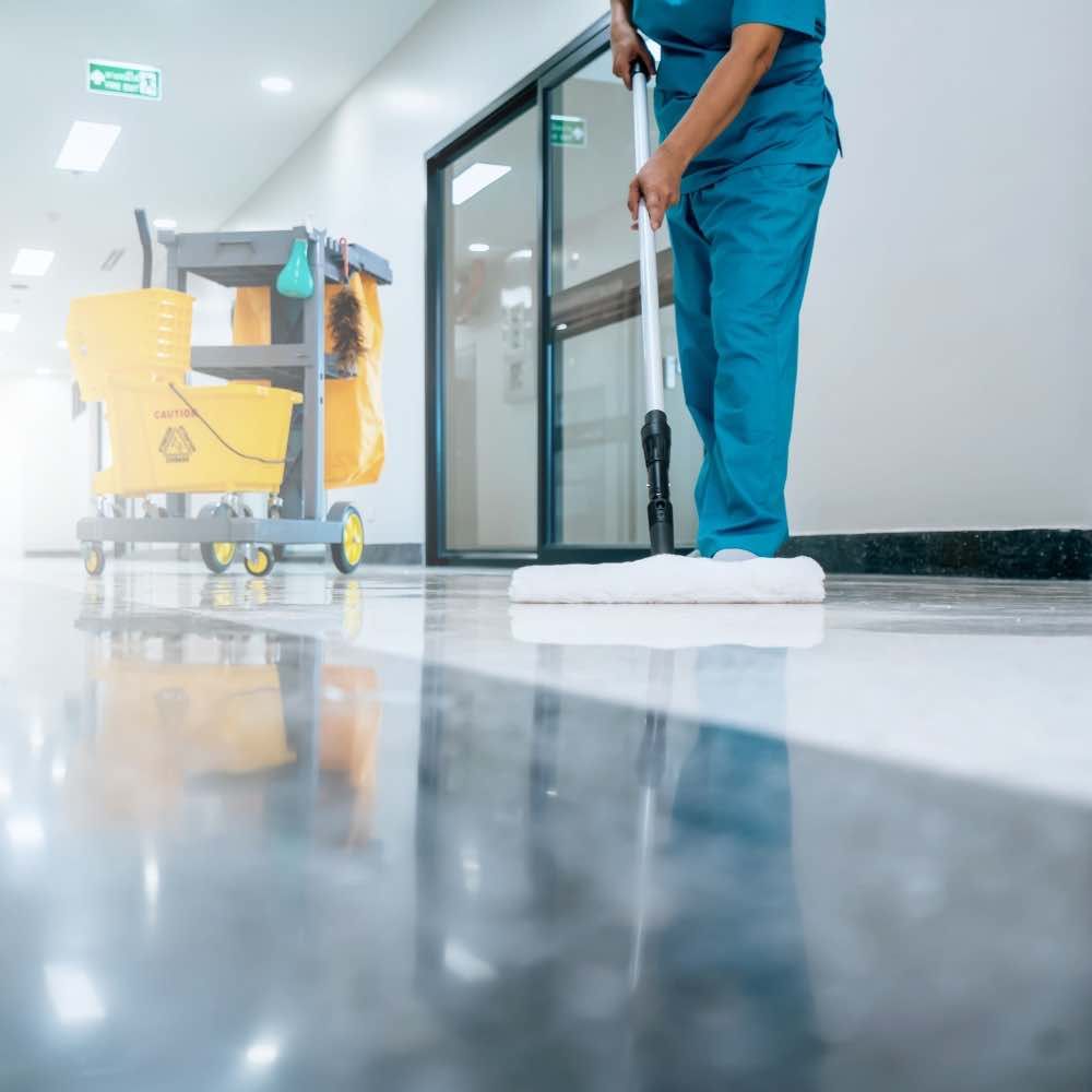 Janitor mopping a glossy floor in a hospital hallway; yellow cleaning supplies cart visible.