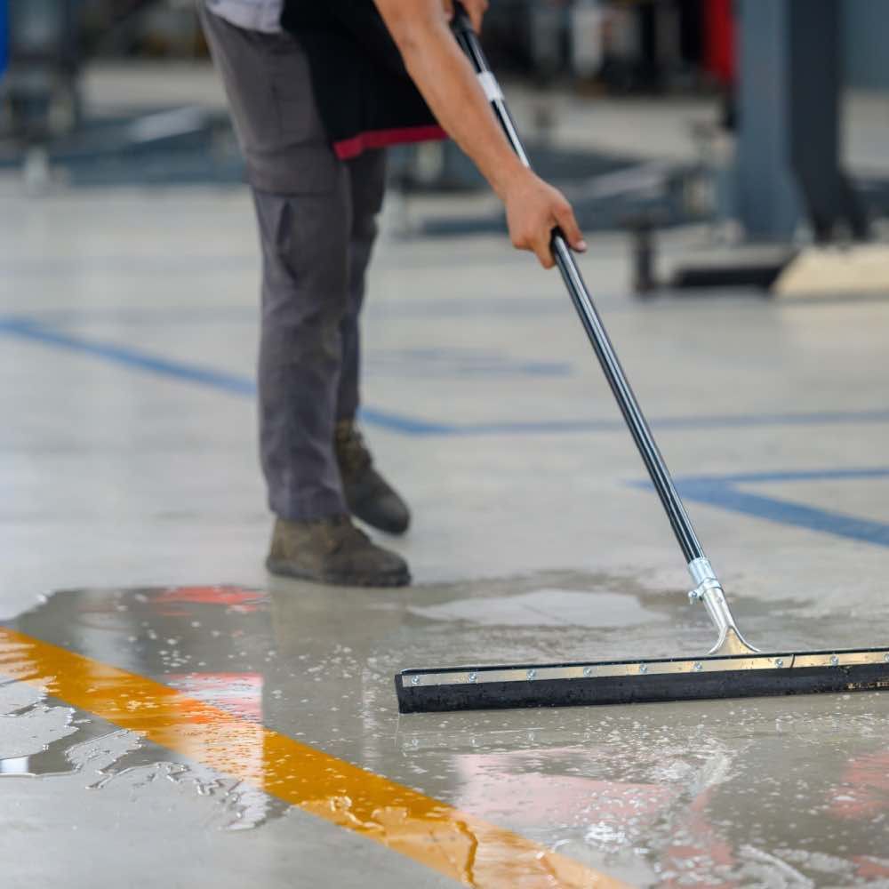 Person squeegeeing a wet floor in a workshop, focusing on cleaning a puddle near a yellow line.