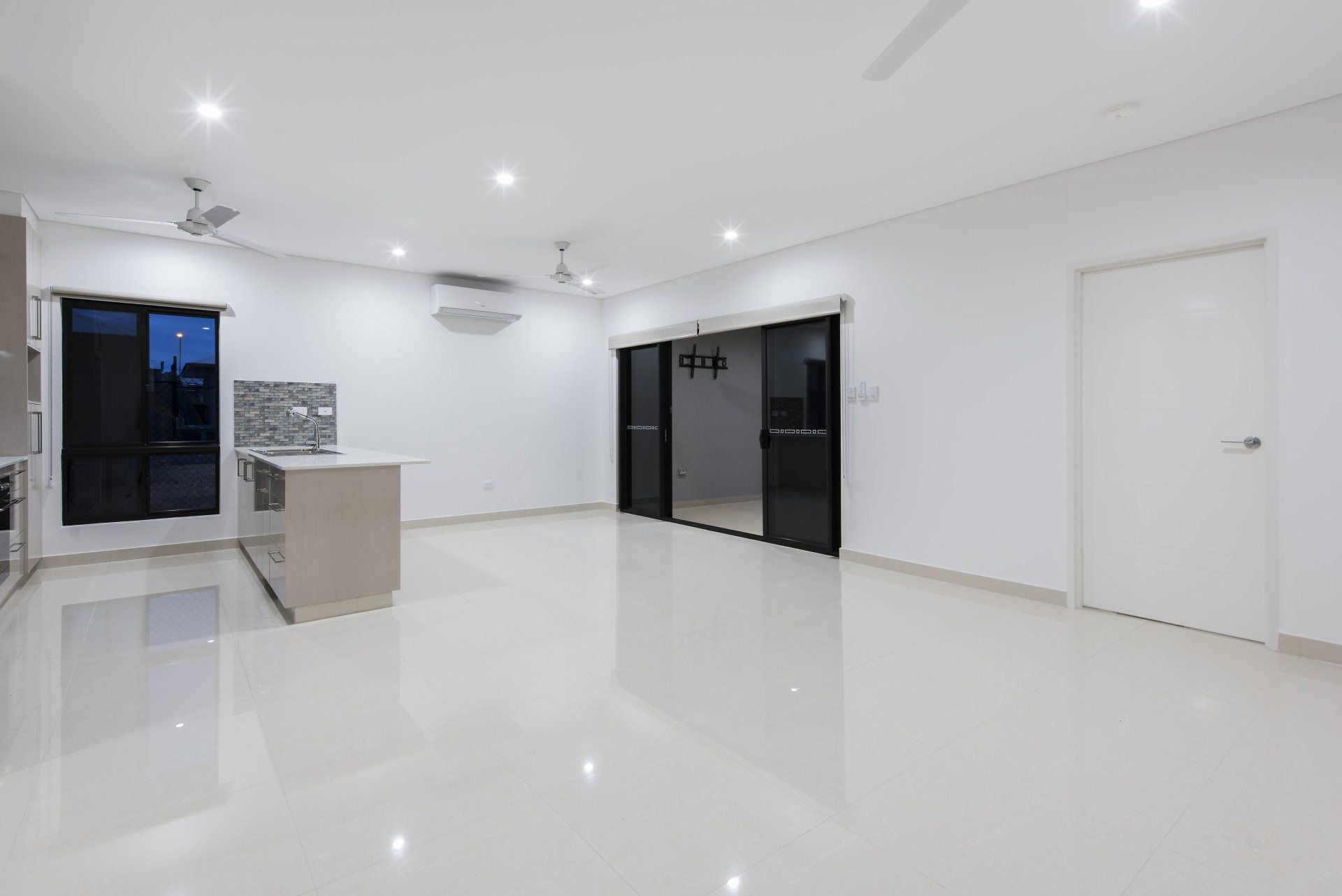 An empty living room with a kitchen and sliding glass doors.