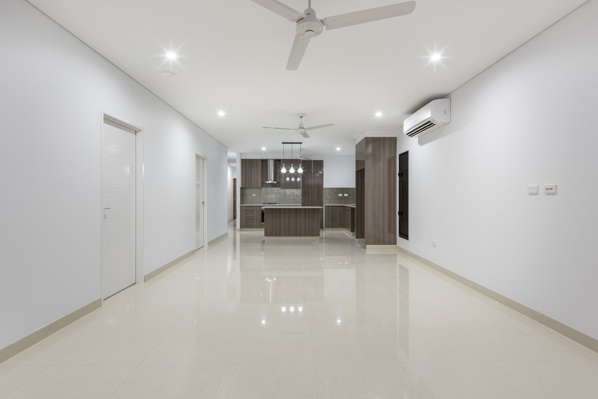 A large empty room with a ceiling fan and a kitchen in the background.