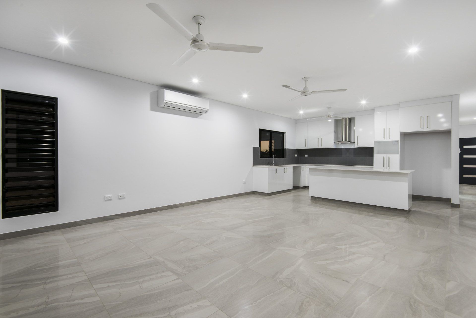 A large empty room with a ceiling fan and a kitchen in the background.